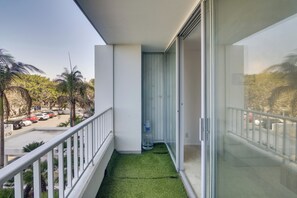 A balcony with a small patch of artificial grass, a glass door, and a view of parked cars and palm trees.