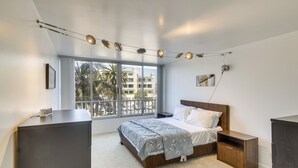 Ein Schlafzimmer mit großem Fenster, einem Bett mit Holz Kopfteil, einem Nachttisch und einem Schreibtisch mit Lampe.