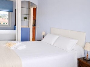 A hotel room with a bed, a nightstand, a lamp, a window, and an open door leading to a kitchen area. - Winahost Piso Gavines (CASTELL-PLATJA D'ARO)