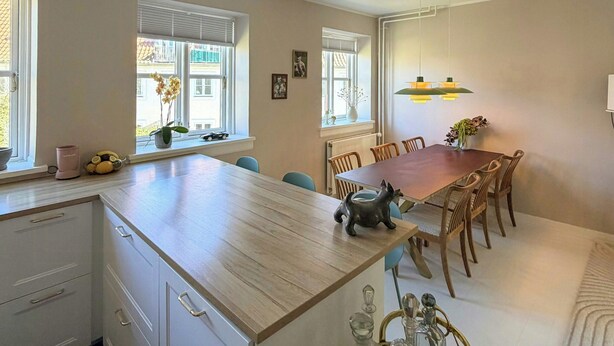 A modern kitchen with a central island, dining table, and chairs. There are windows with blinds, a radiator, and a hanging light fixture. - Light apartment with classic details in Copenhagen (København)