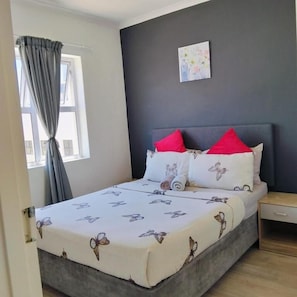 A bedroom with a bed, a nightstand, a window with curtains, and a wall-mounted picture.