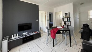 A modern living room with a flat-screen TV, a dining table with chairs, and a kitchen area in the background.