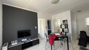A modern living room with a flat-screen TV, a dining table with chairs, and a kitchen area in the background.