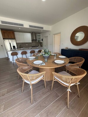 A dining area with a round wooden table surrounded by wicker chairs, a kitchen in the background, and a large mirror on the wall.