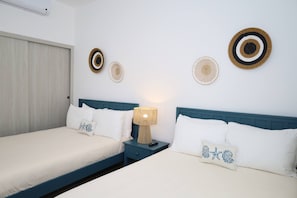 A double bed with white bedding and a blue headboard, a bedside table with a lamp, and decorative woven wall art.