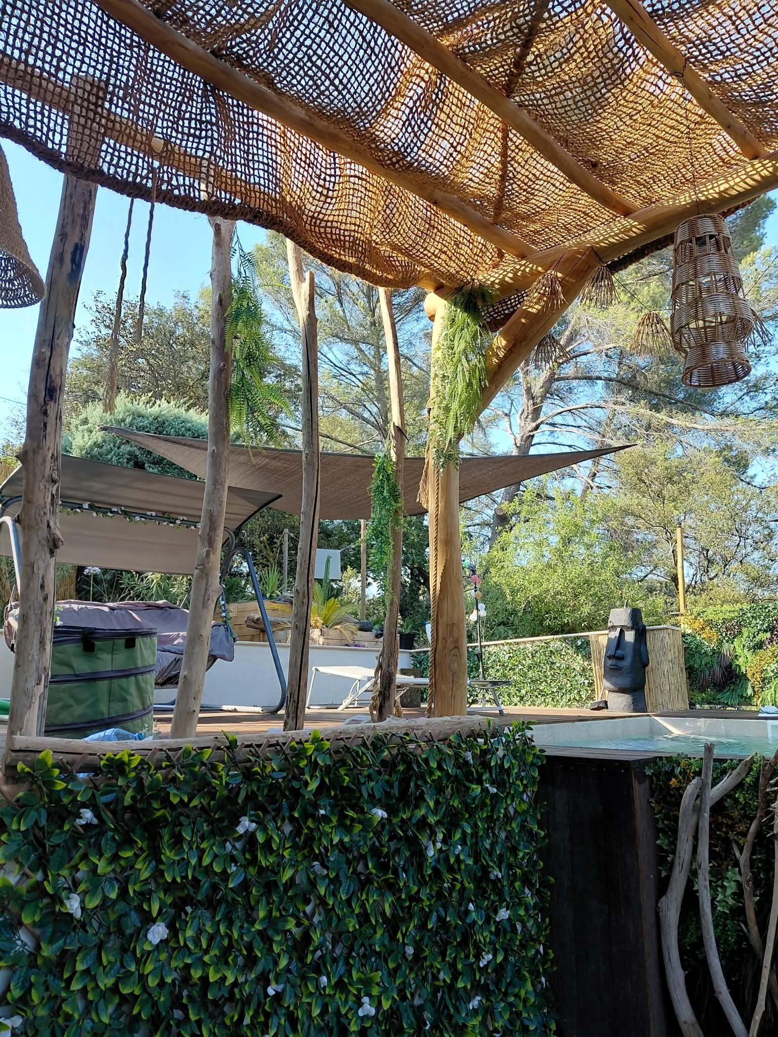 Un espacio al aire libre techado con vigas de madera y techo de malla, rodeado de follaje y árboles.