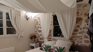 Et soveværelse med stenvæg, en seng med blomstermønstrede puder, et natbord, en lampe og et vindue med gardiner.