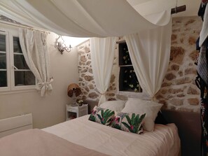 A bedroom with a stone wall, a bed with floral pillows, a nightstand, a lamp, and a window with curtains. - Exceptional, charming home with a private and exclusive pool (La Motte)