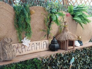 Un mur décoratif agrémenté de paniers tressés, de plantes et d’une enseigne en bois portant le nom « KAZAMIA ».