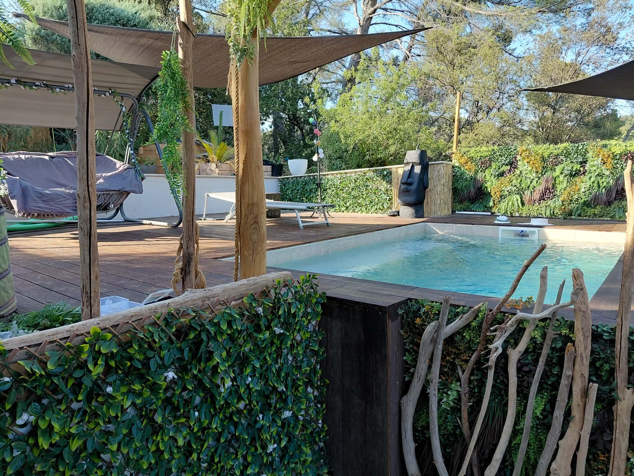 Una piscina amb una terrassa de fusta, envoltada de vegetació i una escultura de cap tiki.