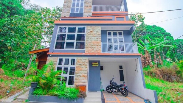 Ein zweistöckiges Gebäude mit einem Balkon im oberen Geschoss, einer Garage mit Motorrad und einem kleinen Garten.