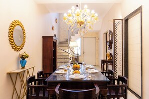 Uma sala de jantar com um candelabro, um espelho com moldura dourada e uma escadaria ao fundo.