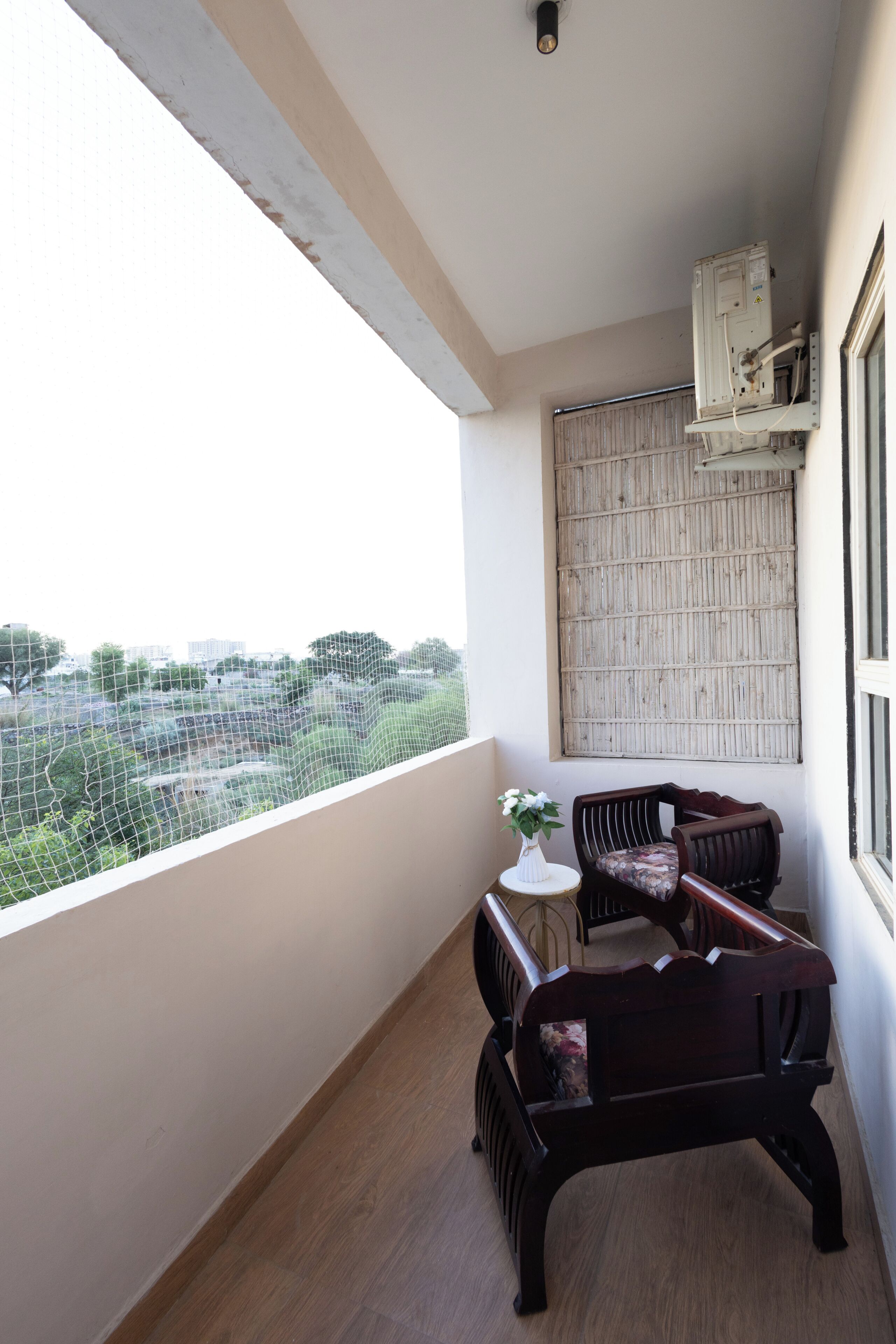 A balcony with a wooden floor, a white table with a vase of flowers, two dark wooden chairs, and a view of a cityscape.