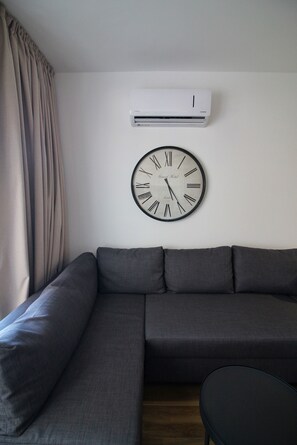 A living room with a sectional sofa, a round wall clock, and an air conditioning unit. - STAY Aspri Petra Apartment (Paphos)