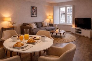 Ein modernes Wohnzimmer mit einem runden Esstisch, Sofas, einem Fernseher und einem Fenster mit Blick auf Gebäude.