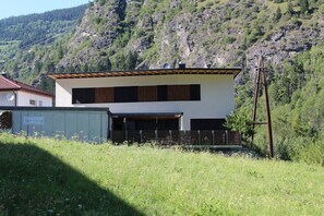 A modern two-story building with a flat roof, large windows, and a garage. It is surrounded by a grassy area with wildflowers and a mountainous backdrop.