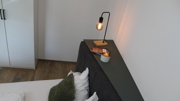 Ein modernes Zimmer mit einem Bett, einem Nachttisch mit Lampe und einem Kleiderschrank.
