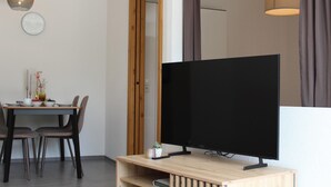 Ein modernes Wohnzimmer mit einem Flachbildfernseher auf einem Holzboden, einem Essbereich mit Tisch und Stühlen sowie einem großen Spiegel, der den Raum reflektiert.