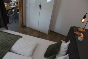 A bedroom with a bed, a nightstand with a lamp, a wardrobe, and a dining area in the background.