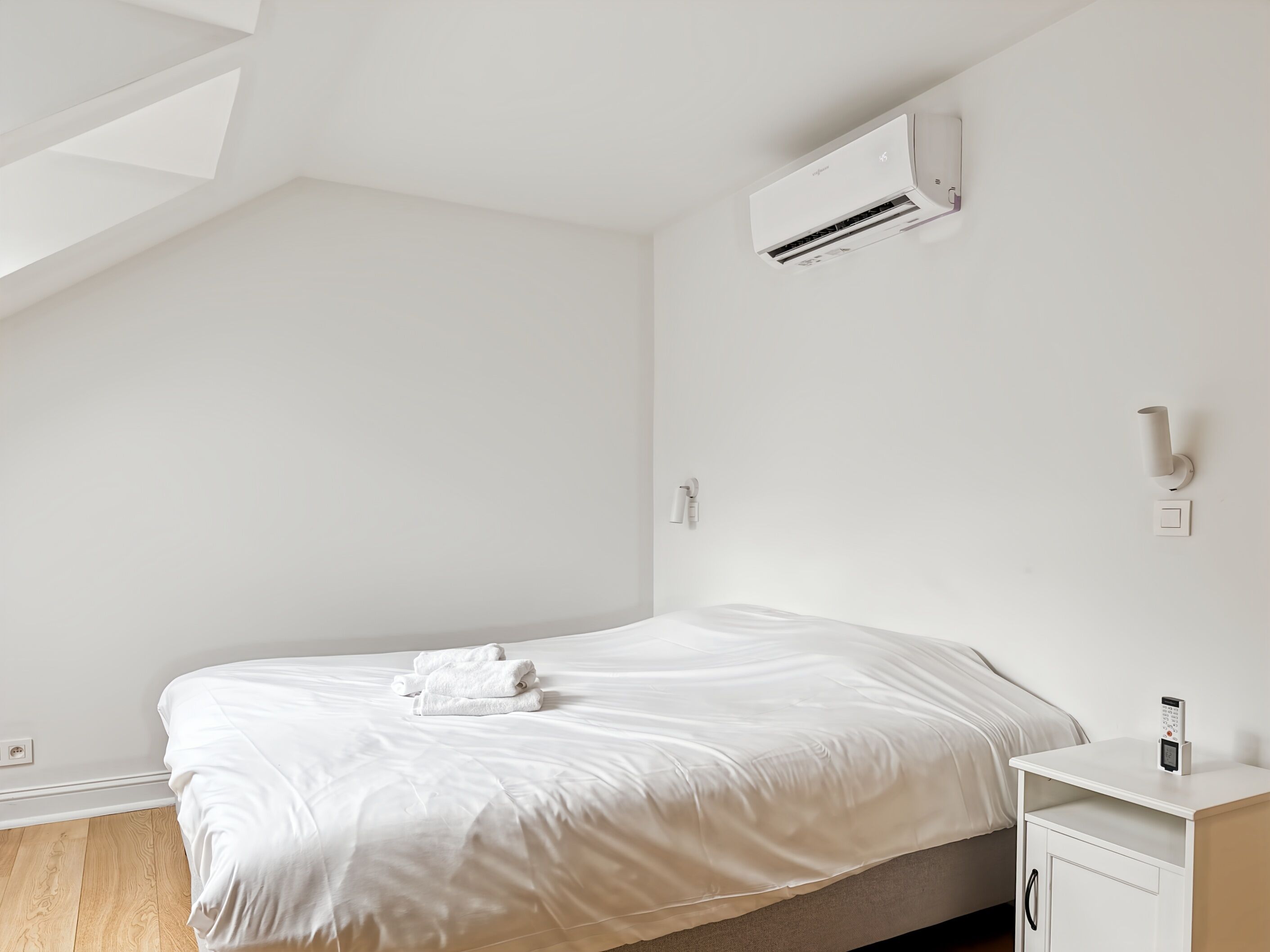 A minimalist bedroom with a bed, a bedside table, an air conditioner, and a wall-mounted light fixture.