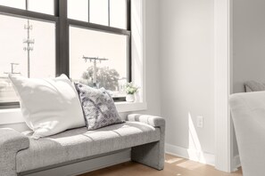 Une chambre moderne avec un banc placé près d’une grande fenêtre, une petite table sur laquelle se trouve un vase, et un lit doté d’une tête de lit blanche.