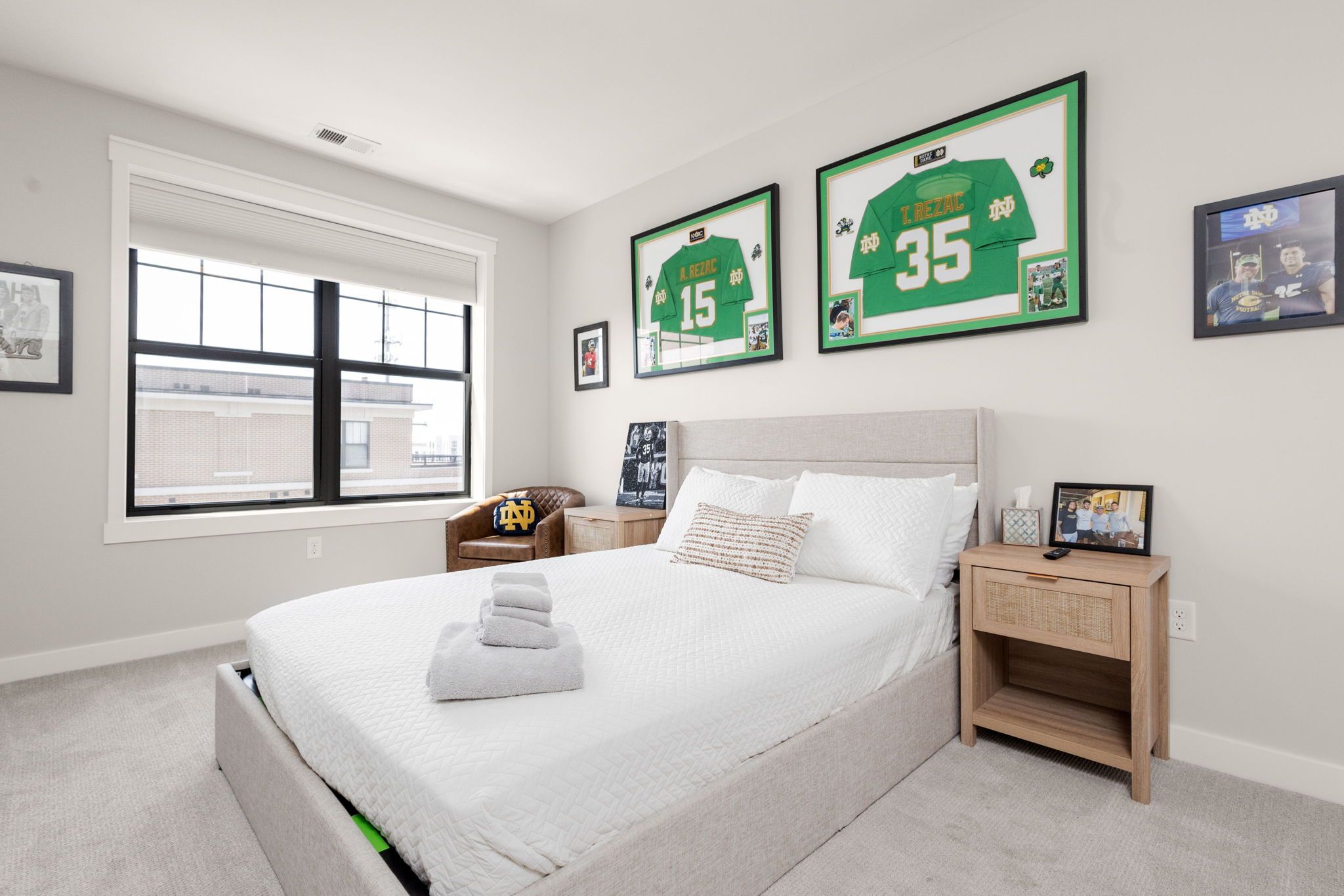 Een slaapkamer met een bed, nachtkastje, raam en ingelijste sportshirts aan de muur.