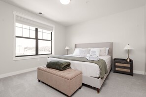 Une chambre à coucher avec un lit, des tables de chevet, une fenêtre et un pouf.