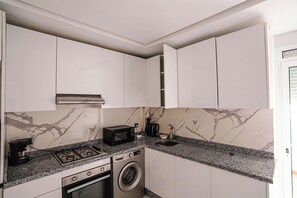 A compact kitchen with white cabinets, a gas stove, a microwave, and a built-in washing machine. - Charming 2-Bedroom Apartment in the Heart of Martil, Morocco (Martil)