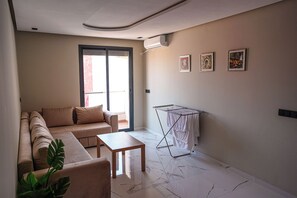 A modern living room with a sofa, a small table, a drying rack, and a sliding glass door leading to an outdoor area. - Charming 2-Bedroom Apartment in the Heart of Martil, Morocco (Martil)