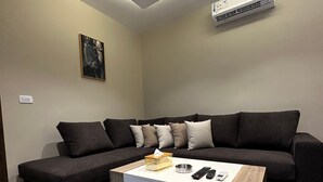 Un salon moderne avec un canapé d’angle, une table basse équipée de télécommandes et un climatiseur fixé au mur.