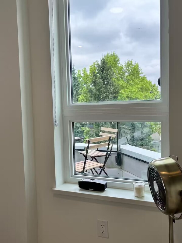 A room with a window overlooking a balcony with a chair and a fan.