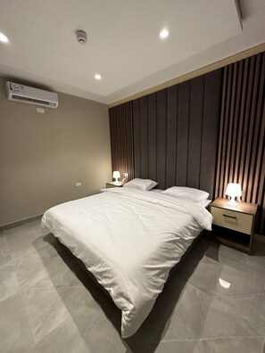 A modern bedroom with a large bed, two bedside tables with lamps, a wall-mounted air conditioner, and a sloped ceiling. - 305 AL-NAJEH JRE Apartments For Rent ave (Amman)
