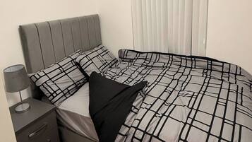 A bedroom with a bed, a nightstand, a lamp, a window with blinds, and a wooden floor.