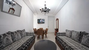 A living room with two patterned sofas, a round coffee table, a dining table with chairs, a TV mounted on the wall, and a chandelier. - Charming 2-Bedroom Apartment in Martil with City Views (Martil)