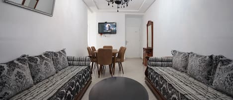 A living room with two patterned sofas, a round coffee table, a dining table with chairs, a TV mounted on the wall, and a chandelier.