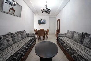 A living room with two patterned sofas, a round coffee table, a dining table with chairs, a TV mounted on the wall, and a chandelier. - Charming 2-Bedroom Apartment in Martil with City Views (Martil)