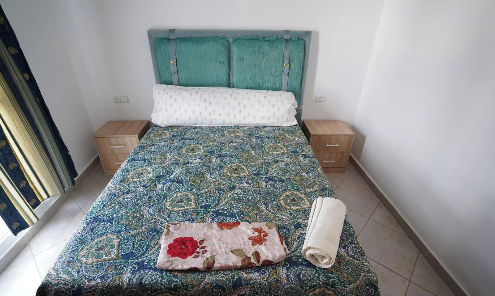 A bed with a patterned bedspread, a pillow with a floral design, and a rolled towel. There are two bedside tables with drawers.