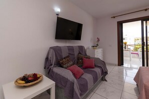 Ein Wohnzimmer mit einer Couch, einem Flachbildfernseher, einem kleinen Tisch mit Obstkorb und einem Balkon mit Aussicht.