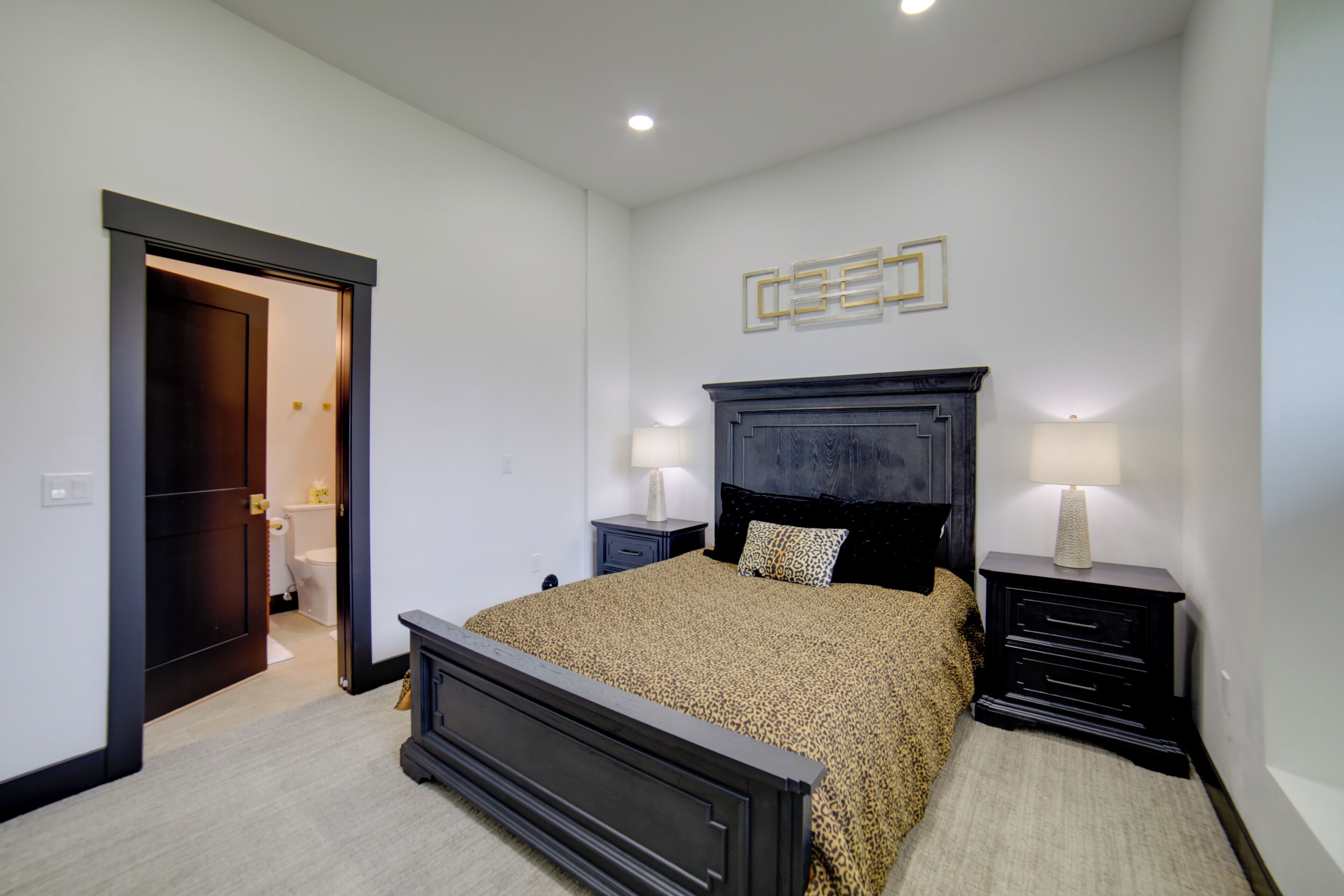 Un dormitorio con una cama grande, mesitas de noche, una mesita auxiliar, una puerta que da al baño y una lámpara fijada a la pared.
