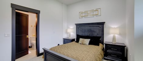 Un dormitorio con una cama grande, mesitas de noche, una mesita auxiliar, una puerta que da al baño y una lámpara fijada a la pared.