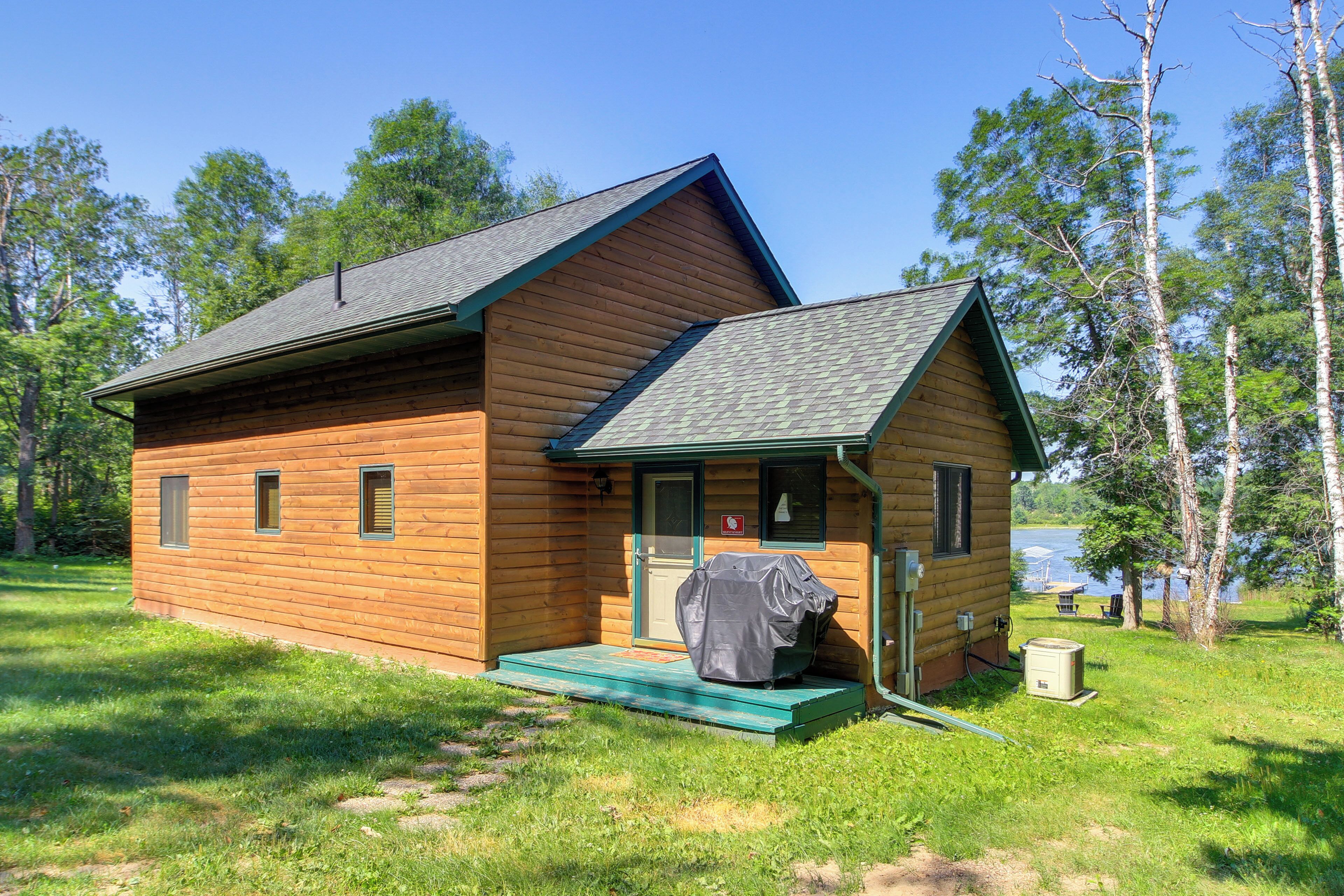 A small wooden cabin with a porch, a covered grill, and a view of a lake.