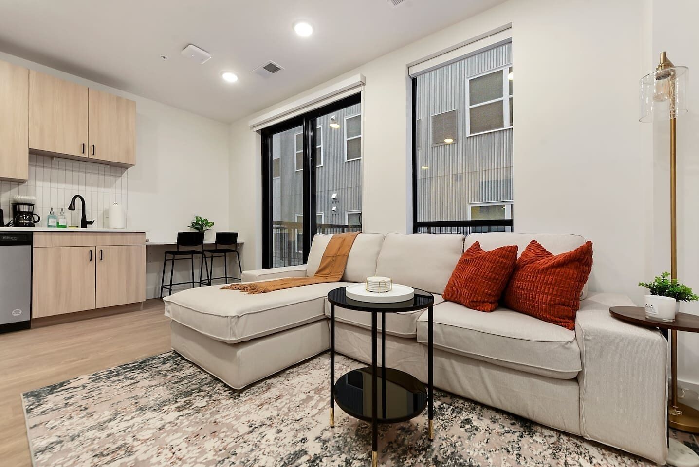 A modern living room with a sectional sofa, a small round table, and a kitchen area with wooden cabinets and a sink.