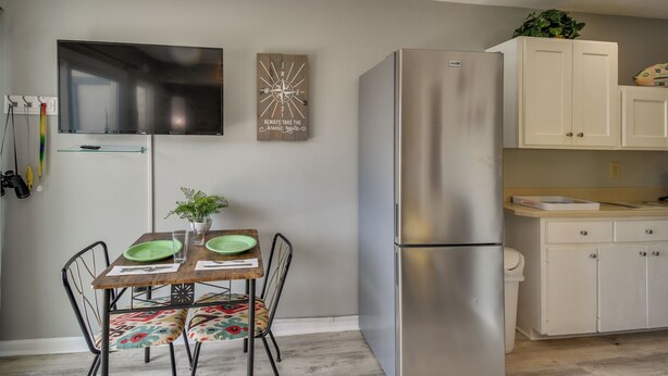 A modern kitchen with a stainless steel refrigerator, a dining table with chairs, and a wall-mounted flat-screen TV.