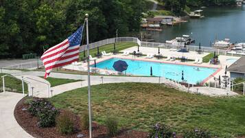 A swimming pool with a flag, surrounded by a deck with chairs and umbrellas, and a lake in the background.