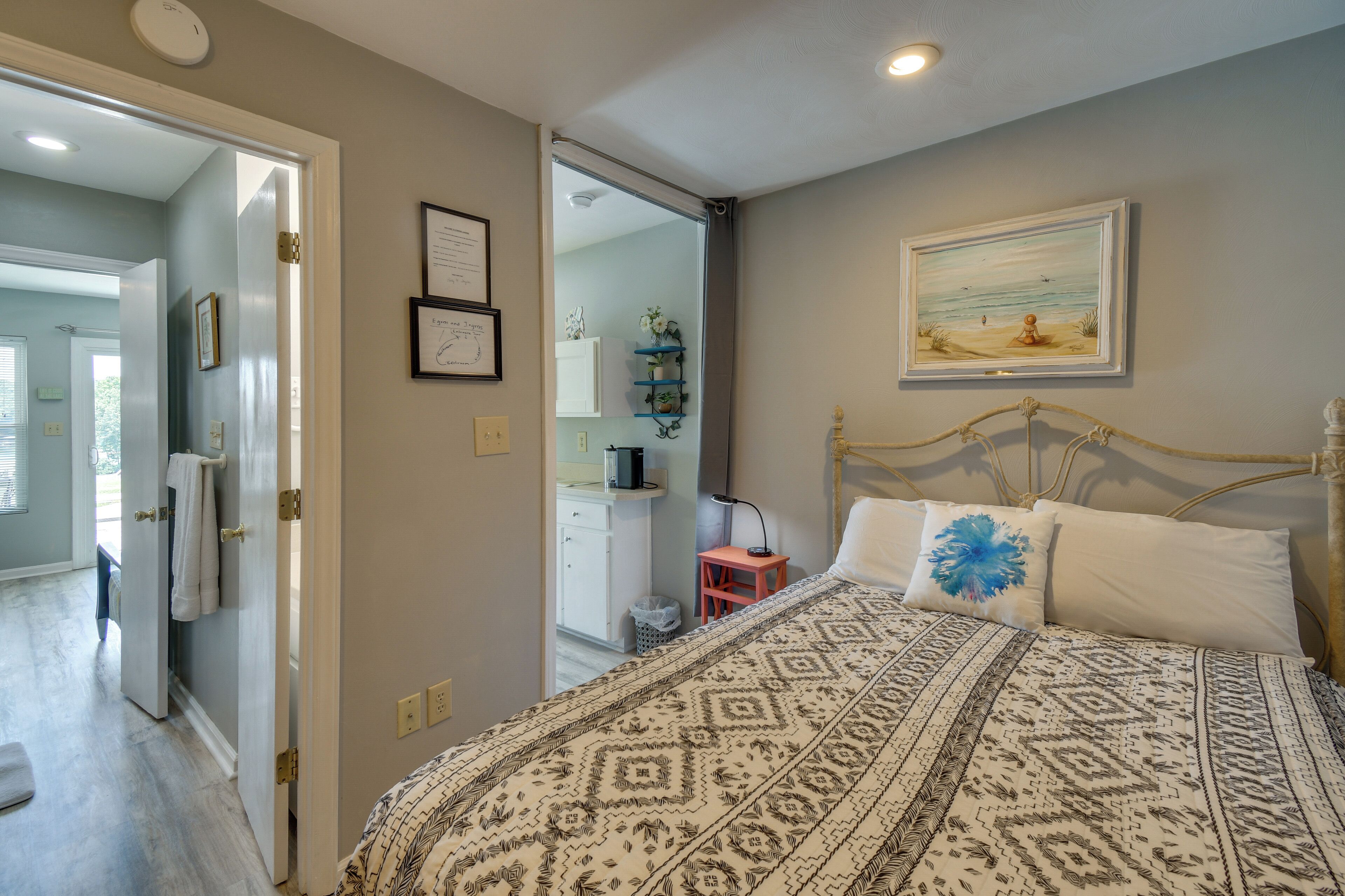 A bedroom with a bed, a nightstand, a painting, and a bathroom visible through an open door.