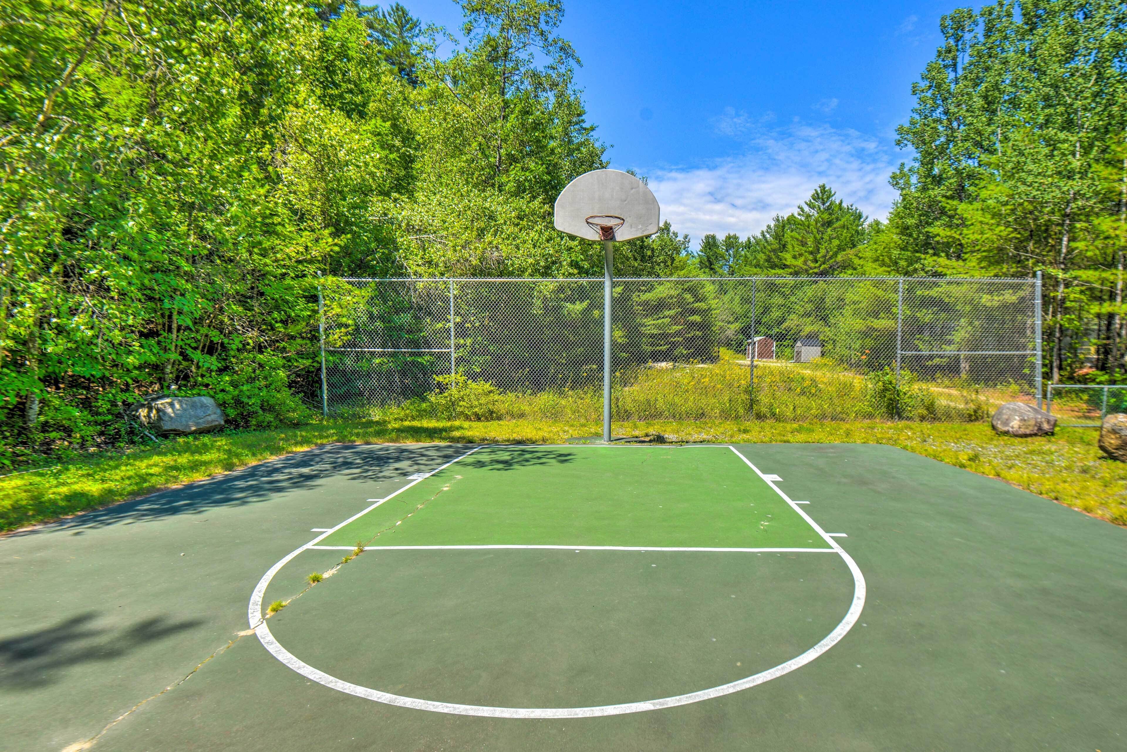 An outdoor basketball court with a chain-link fence, surrounded by trees and a clear sky.