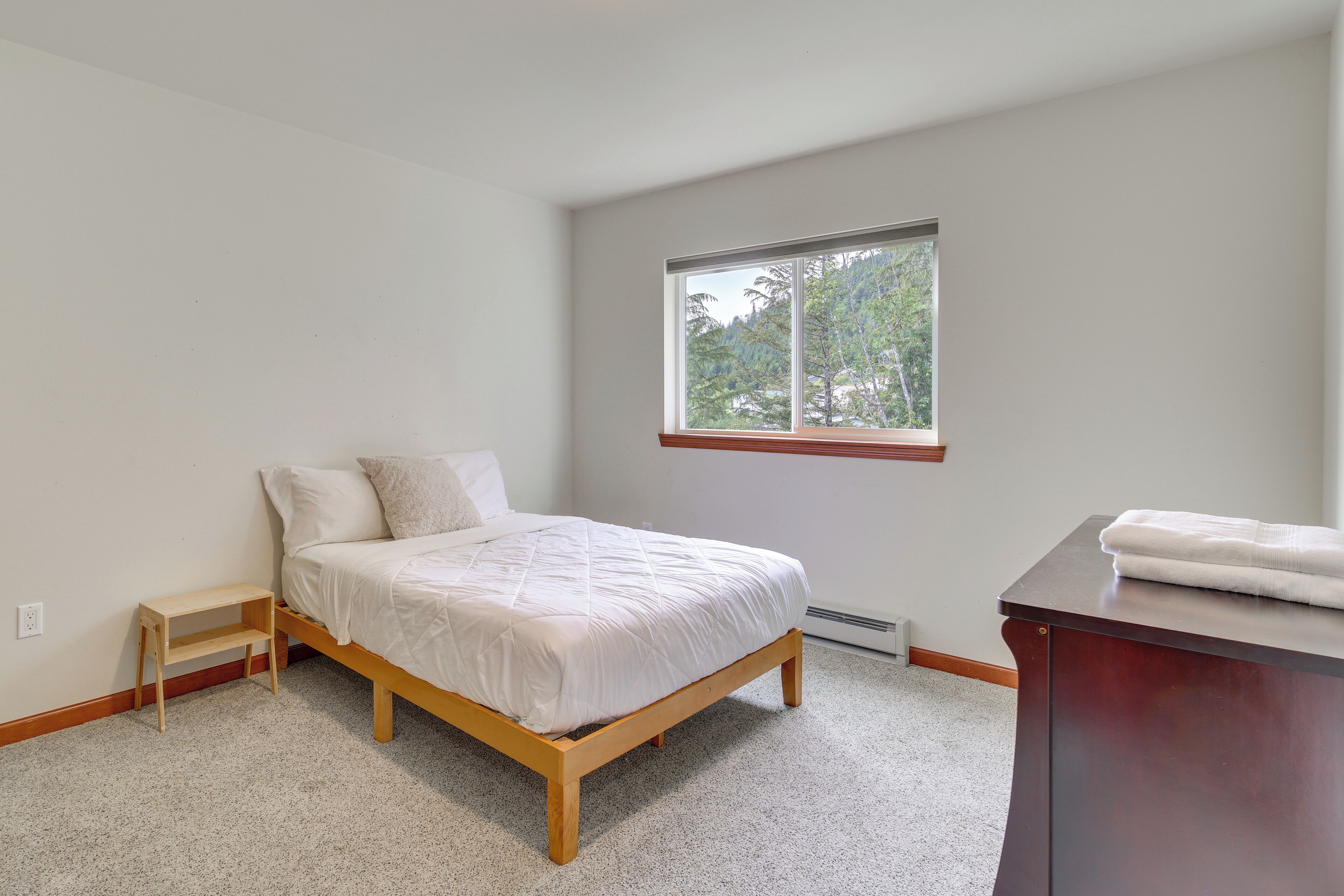 A bedroom with a bed, a small wooden nightstand, a window with a view of trees, and a dark wooden dresser.