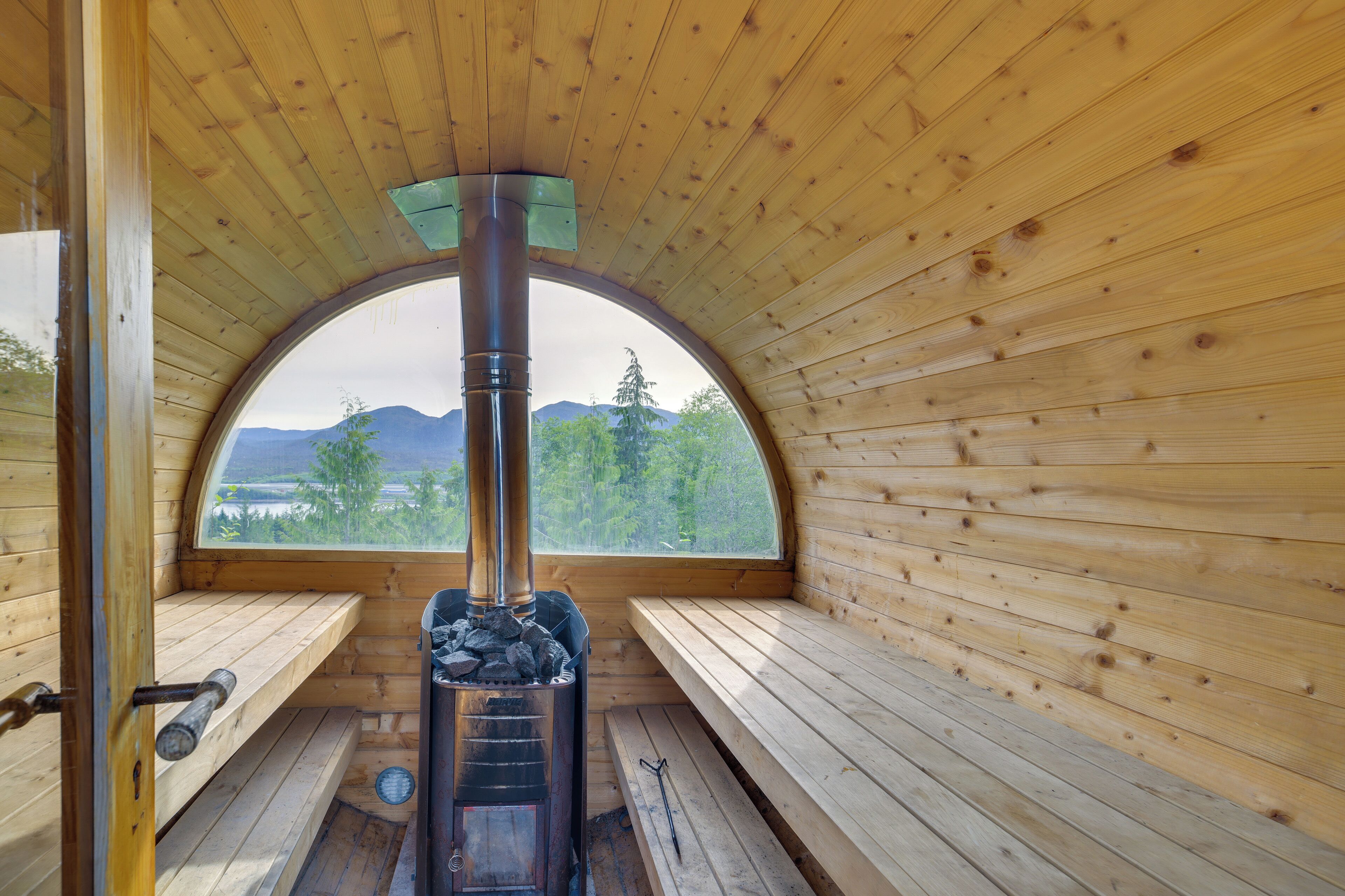 A wooden cabin with a central fireplace, benches, and a view of mountains and trees through a large arched window.