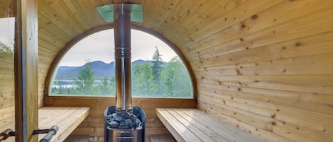 A wooden cabin with a central fireplace, benches, and a view of mountains and trees through a large arched window.