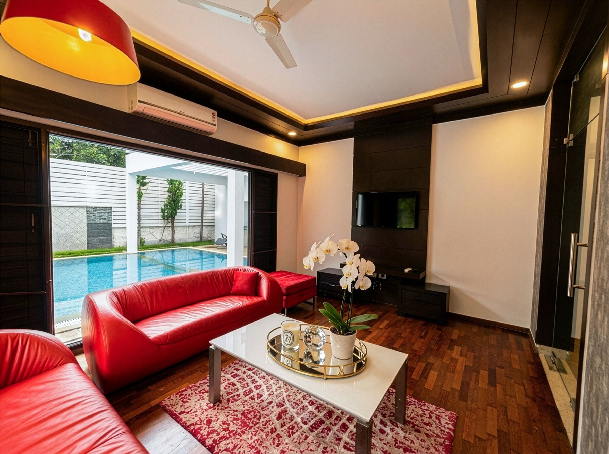 Uma sala de estar moderna com um sofá vermelho, uma mesa de centro branca e uma televisão. É visível uma piscina através da porta de vidro.
